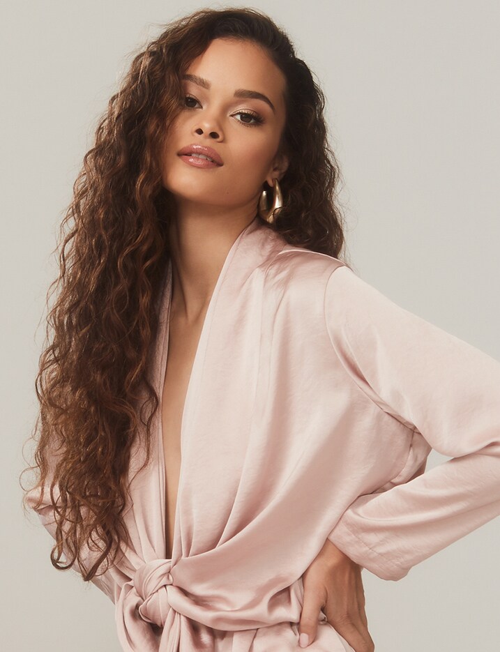 A model with long curly brown hair has her hands on her hips wearing a light pink blouse. Shop Hair.