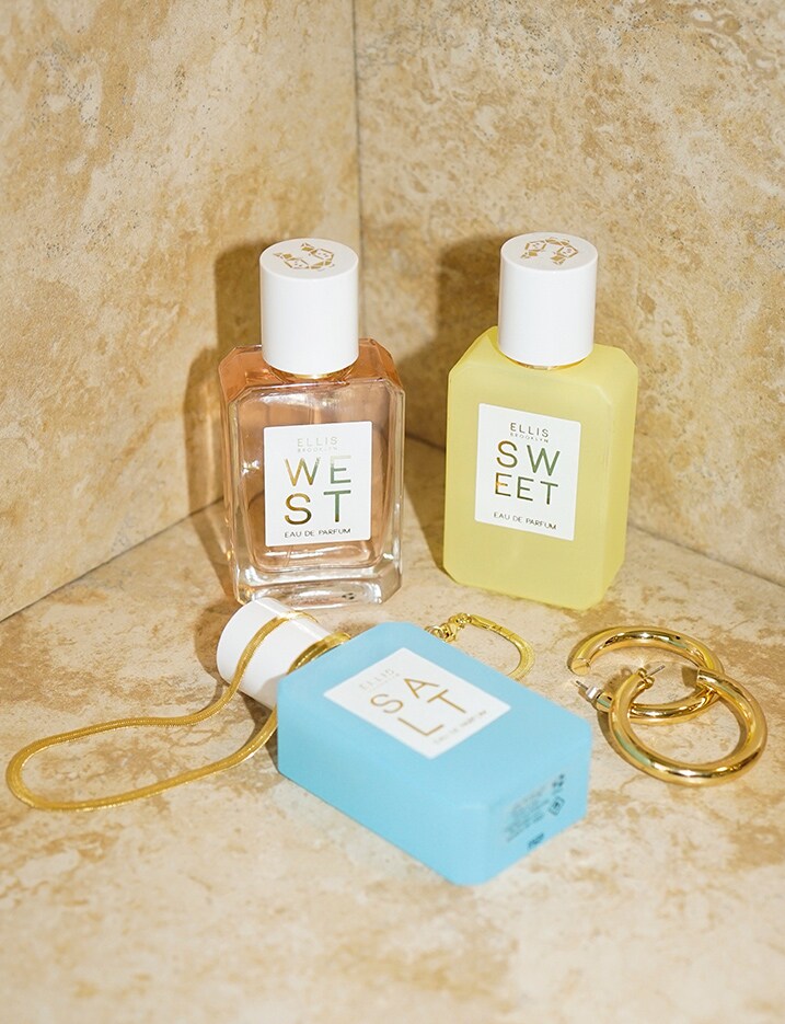 A pink bottle labeled WEST, yellow bottle labeled SWEET and blue bottle labeled SALT from Ellis Brooklyn Eau de Perfume sit on stone tiles. Shop Fragrance.