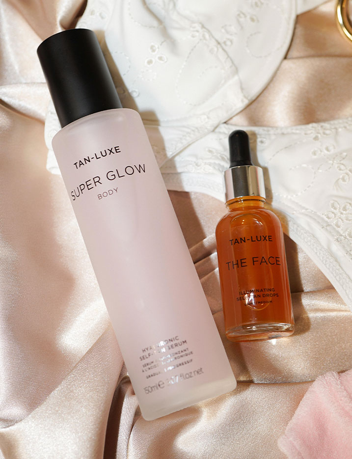 Tan-Luxe self-tan serum and illuminating self-tan drops sit on satin and lace fabric. Shop Self Tanners.