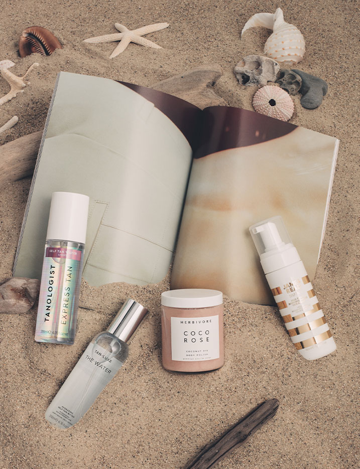 A variety of self-tanners and tanning skincare products sit in the sand with a magazine and seashells. Shop Beach-Ready Bronze.
