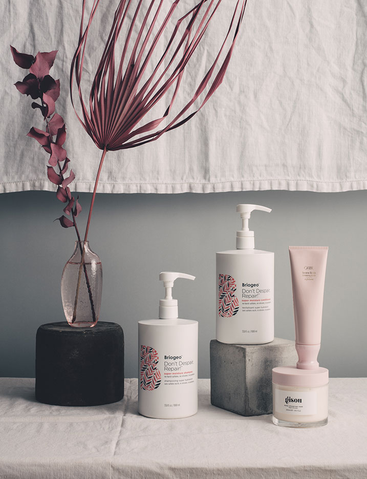 Haircare products are displayed on a canvas covered table cloth with concrete stones and rocks and a vase of burgundy fronds. Shop Hair Game-Changers.