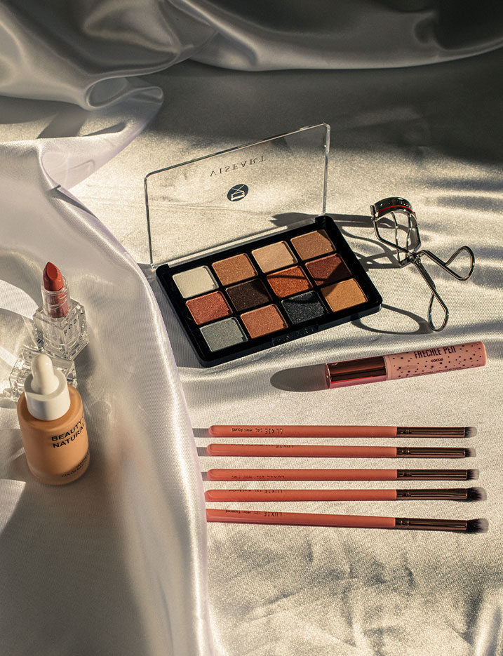 Eyeshadow palette, foundation, lipstick and makeup tools scattered on satin sheet.