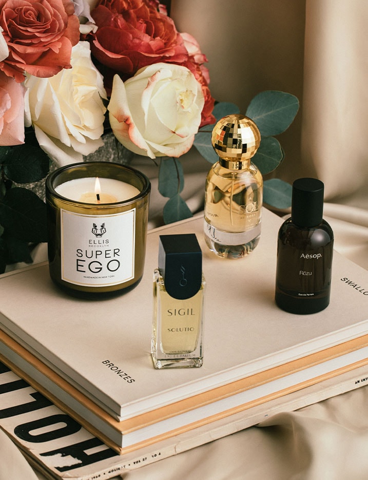 A variety of fragrances in candle and bottled oil form sit on a table of roses and books. Shop Fragrance.