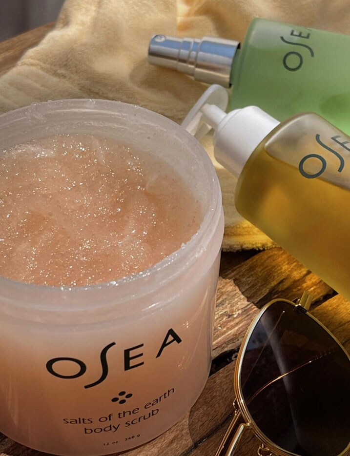 Osea brand bath and body salt scrubs, oils and mists lay on a towel with sunglasses.