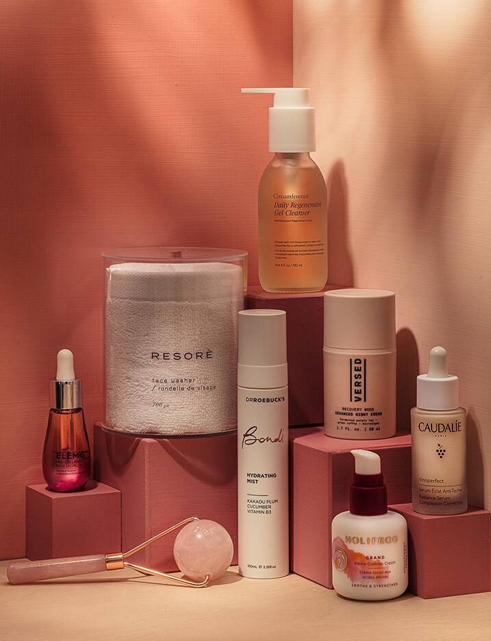 A variety of skincare creams, serums, and oils are displayed upon tiers of pink boxes.