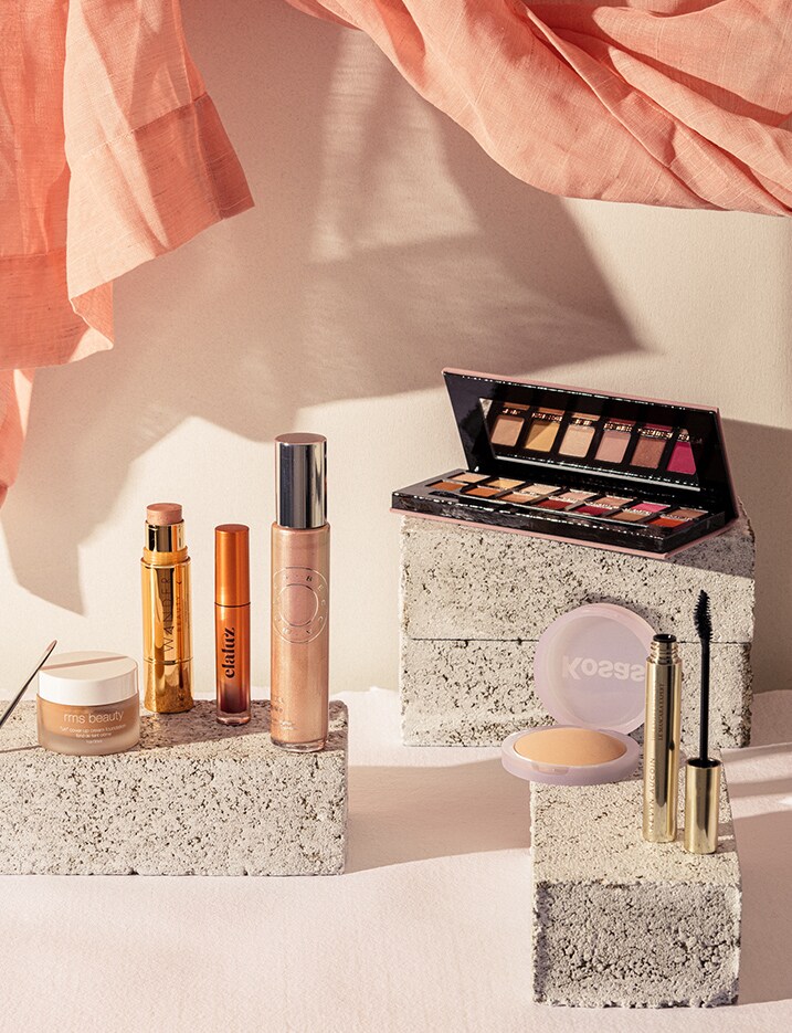 A variety of makeup products sit on stone blocks with a peach-toned linen drape floating lightly above. Shop Makeup.