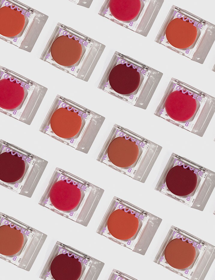 A variety of shades of tinted balms packaged in square, clear lucite compacts are displayed neatly in a diagonal grid on a white surface.