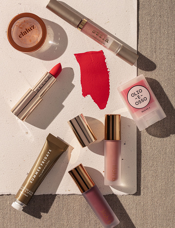 A variety of lip sticks, lip balms and lip oils lay scatered on textured paper and linen.