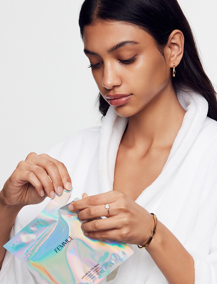 Model dressed in white bath robe opens up an irridescent package of a sheet mask from Femmue brand.
