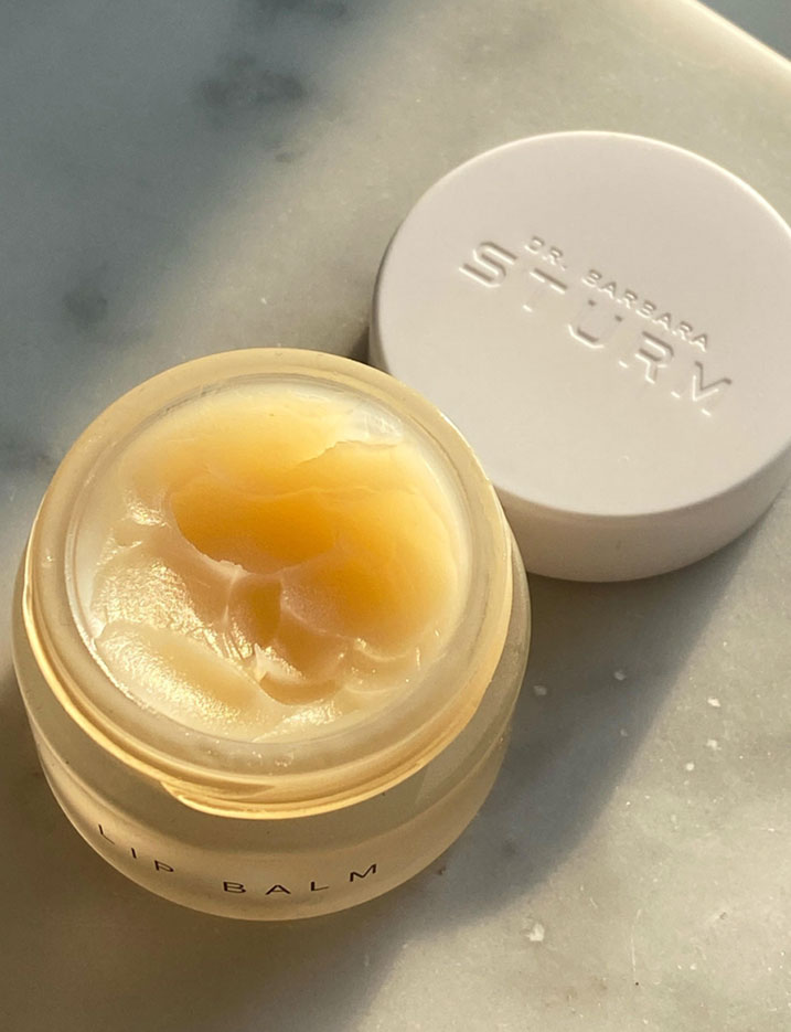 A small opened glass pot of Dr. Barbara Sturm Lip Balm reveals smooth divots in the thick balm.