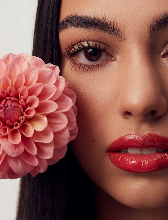 Model holds up a large pink zinnia flower to her face. She is wearing a soft bright geranium red colored lipstick.