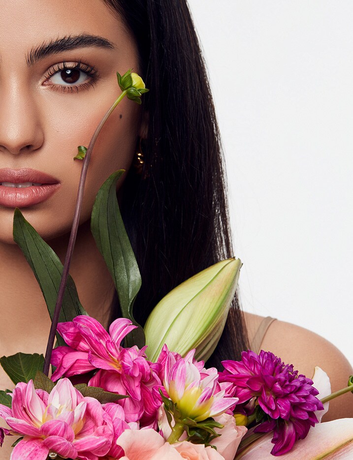 Model with full dark brows holding a bouquet of flowers below her chin.