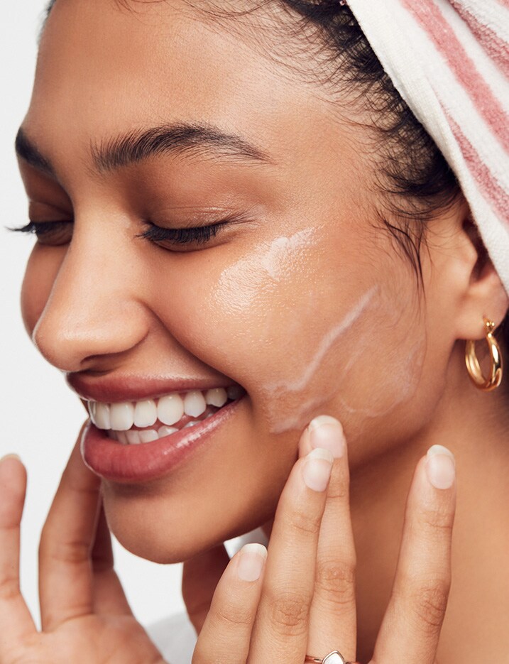 Model smiling with her eyes closed as she gently lathers face wash onto her cheeks with her fingertips.