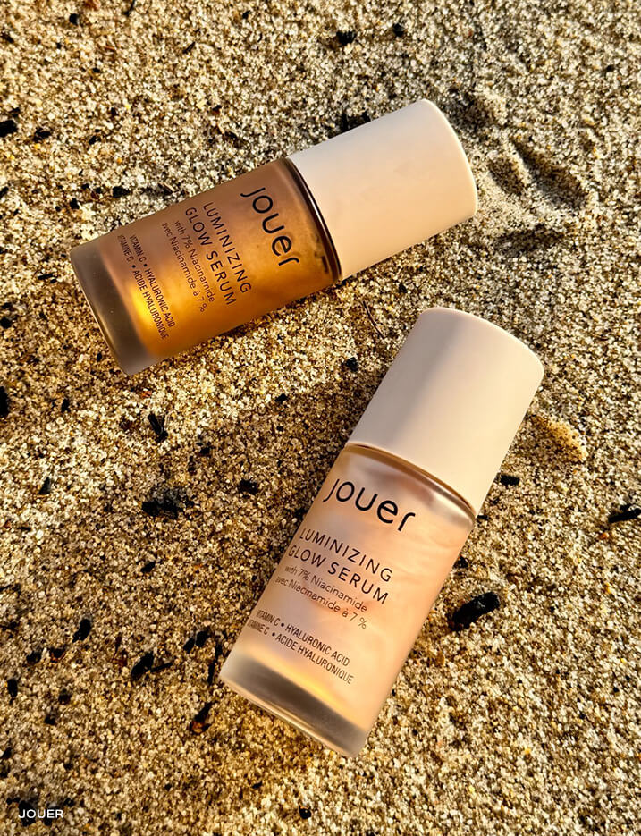 Bottles of Jouer Luminizing Glow Serum on the sand.
