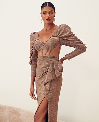 A model standing with one arm on her hip wearing a gold shimmer long sleeve gown with front panel cut-outs and puff sleeves. Enter the Holiday Shop.