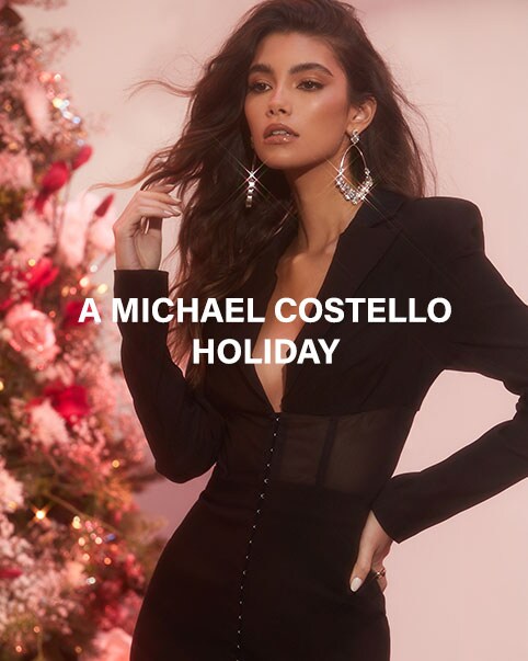 A model wearing a one sleeve black dress holding a present in front of a tree decorated with lights and flowers. A model standing in front of a tree decorated with lights and flowers wearing a black blazer dress with padded shoulders, a hook and eye front, a semi-sheer waist, with silver drop earrings. A Michael Costello Holiday. Shop the Collection.