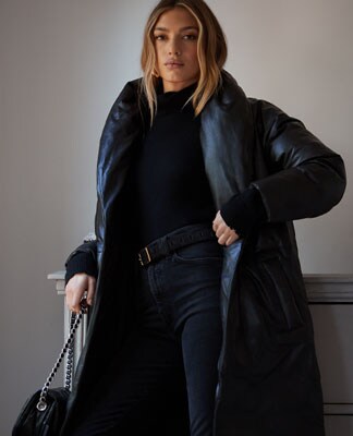 A model standing in front of a chest of drawers wearing a black mock neck sweater, a long black puffer coat, black jeans, and black boots. Coat Check. Shop the Outerwear Edit.