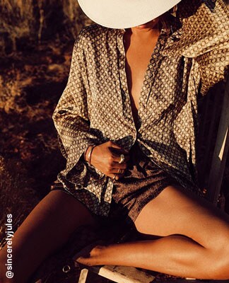 Julie Sari&ntilde;ana sitting outside on a chair wearing a tan colored patterned long sleeve button down top and matching shorts and a white hat. The Perfect Pairings. Shop Now.