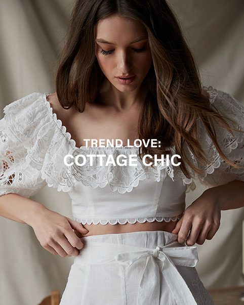 A model sitting in front of a chair wearing a yellow, pink, and blue maxi dress with puff sleeves and eyelet detail. A model wearing a white copy top with lace, ruffle, and scallop details, and white high waisted shorts with a waist tie. A model standing in front of a table and chair wearing a yellow, pink, and blue maxi dress with puff sleeves and eyelet detail, and holding a straw bag. A model wearing a pink cardigan with floral embroidery and cream colored belted high waist pants. Trend Love: Cottage Chic. Shop the Edit.