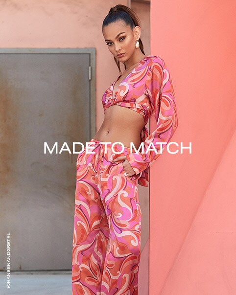 Three photos of a model wearing a hot pink silky matching set that has a swirl pattern all over in shades of cream, purple, pink, and orange. The set features a long sleeve crop top that has a deep v neckline and a crisscross waist that ties in the back and a pair of low-rise pants. Made to Match. Shop Matching Sets.