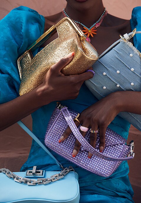 Model holding a gold clutch, a light blue shoulder bag detailed with pearls, and a purple studded ba