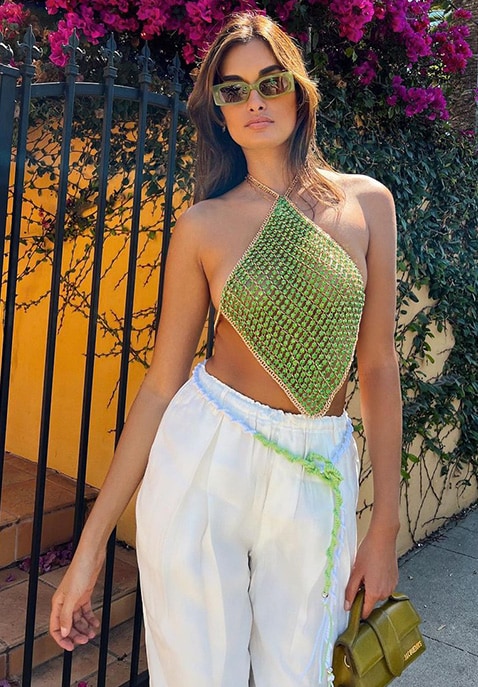 Model in a green beaded halter style top and white pants stands against a color backdrop of pink flo