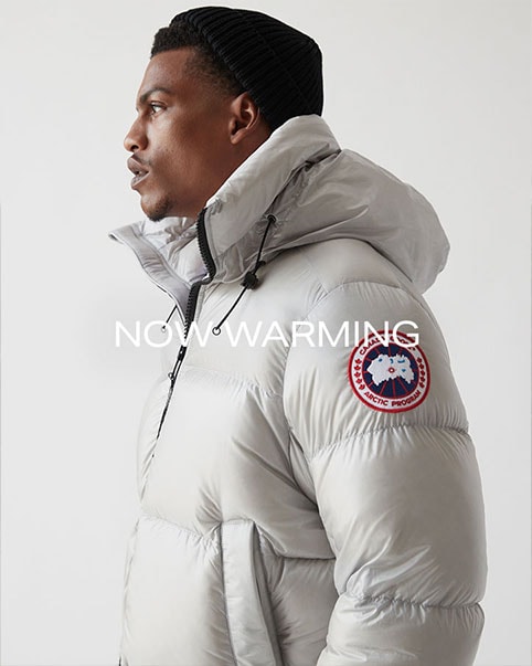Close up image of model’s profile wearing a Canada Goose white hooded jacket and black beanie next to a full body image of the same look. The model is wearing a white shirt under a black sweatshirt with black sweatpants and black boots. Now Warming. Keep the cold away with these superior outerwear styles. SHOP OUTERWEAR