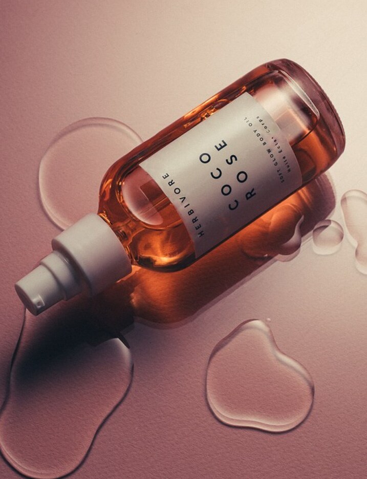 A bottle of Coco Rose body oil from Herbivore lays on its side over appearing to drip in a puddle of oil.
