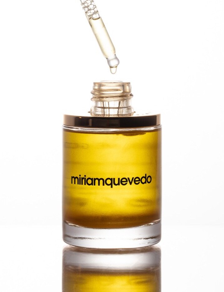 An oil dropper with a drip of nourishing hair oil hovers over a glass bottle of a gold color oil from Miriam Quevedo.
