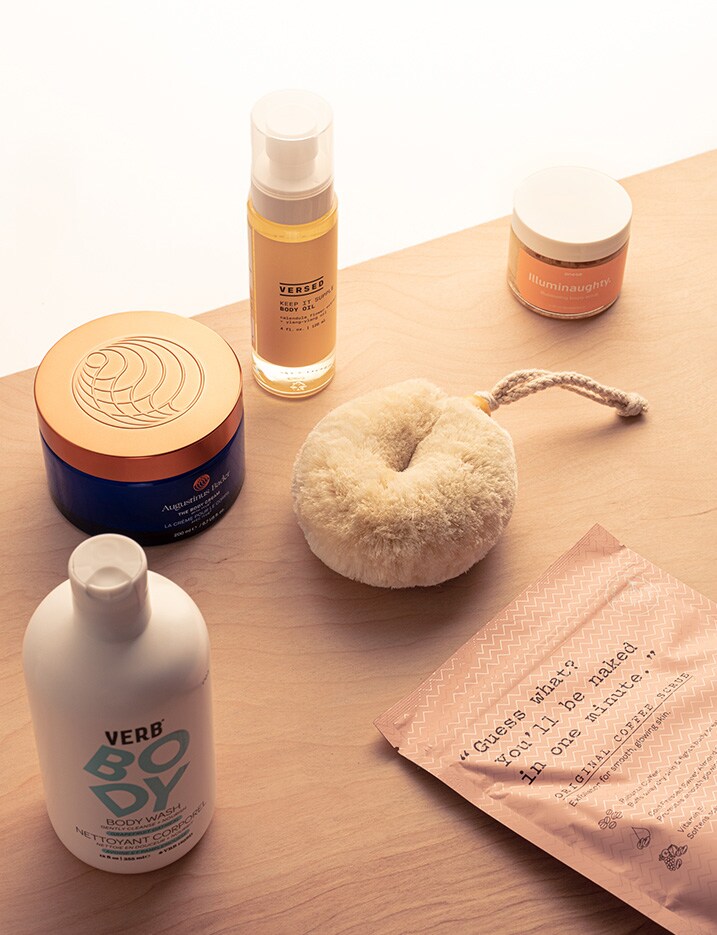 Body skincare products such as natural loofa, scrubs, creams, oil, and lotions sit on a wooden surface.