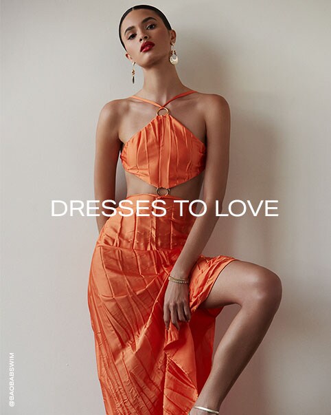 Two photos of a model wearing a slightly pleated orange maxi dress that features a handkerchief top with a halter tie and a silver ring connecting the halter tie to the top and another silver ring connecting the top to the skirt, which creates two large side cutouts. Dresses to Love. Enter the Dress Shop.