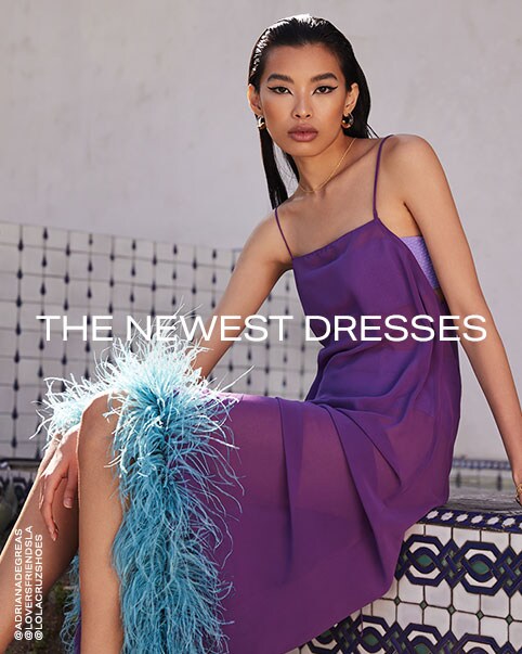 Two photos of a model wearing a bright purple maxi dress that features a squared off neckline and an open back and is slightly see-through. The dress also has bright blue feathers all along the hem. The skirt of the dress is slightly higher in the front than the back. She is wearing a lilac strapless bikini top and high-waisted bikini bottoms underneath. She also has it paired with a pair of metallic light purple strappy heels. The Newest Dresses. Shop New Dresses.