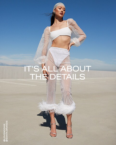Two photos of a model wearing a white long-sleeved crop top. The sleeves of the top are sheer & gauzey & feature a slight puff to them. She has it paired with a pair of white mesh rhinestone pants worn over a pair of white bikini bottoms. The pants have feathers around the ankle. She is also wearing a white mesh rhinestone hair tie that goes over the entire top of her head and a pair of silver heels. It’s All About the Details. Shop the Edit.
