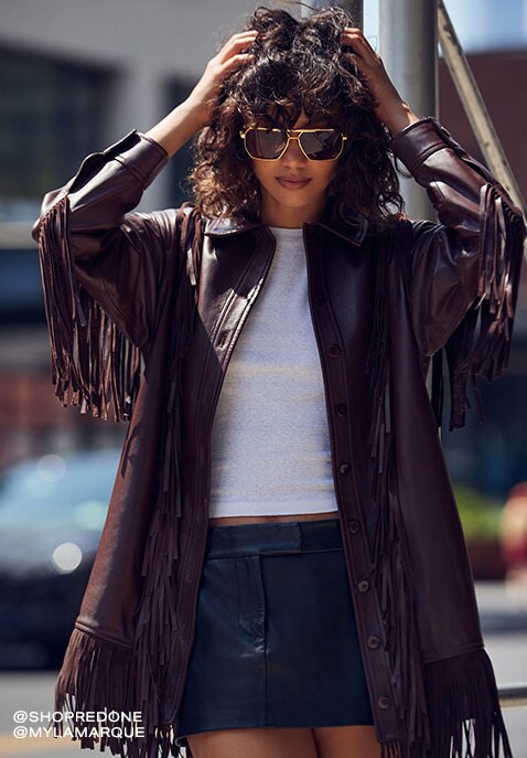 Model wearing a fringed dark maroon jacket over a white tee and a black mini skirt runs her fingers 