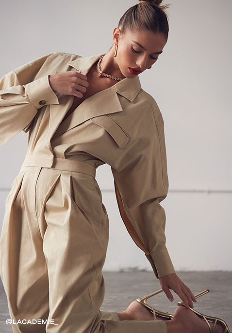Model wearing a khaki jumpsuit with exaggerated sleeves leans back on her knees reaching towards the