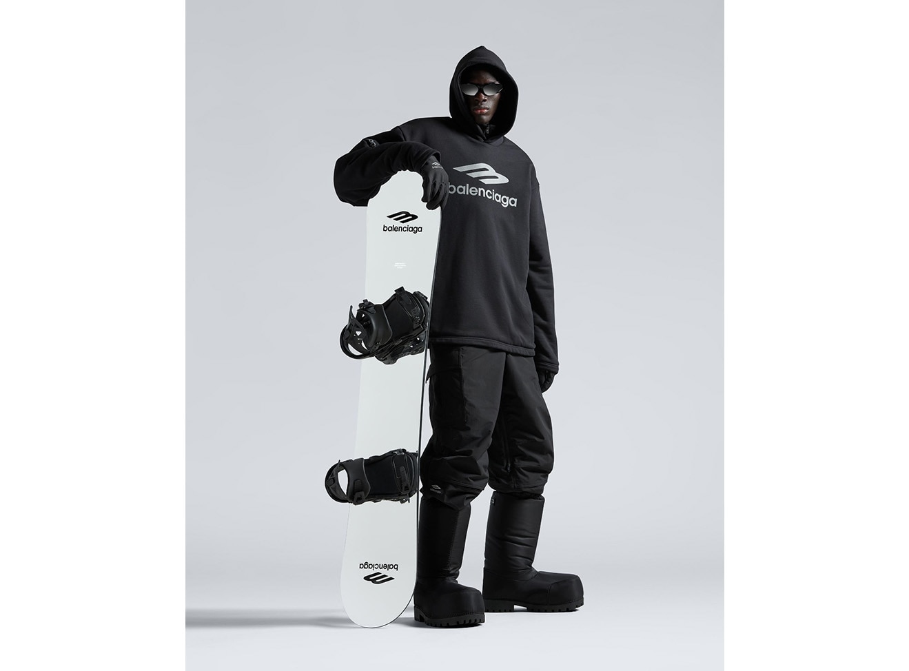 A close up of a model in all black Balenciaga snow gear holding a snowboard. 