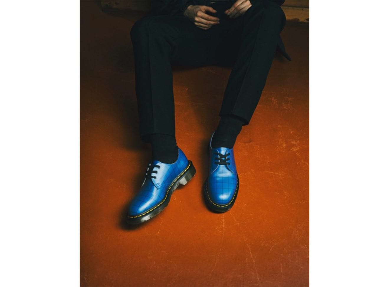 Model sitting down wearing a blue patent leather Dr. Marten x Undercover shoe.