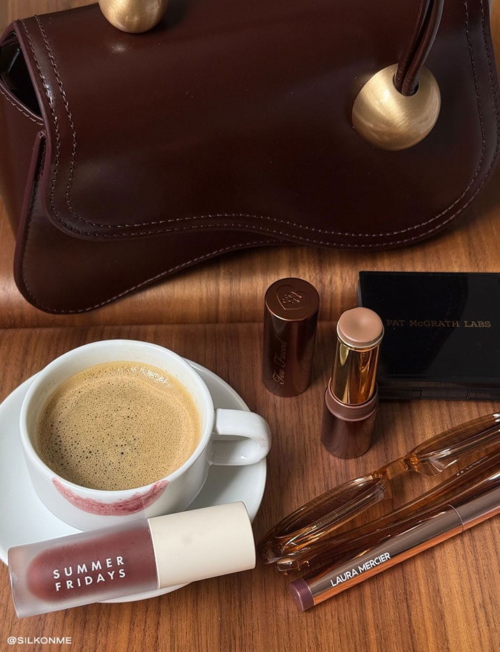 Variety of makeup products next to a cup of coffee and a purse.