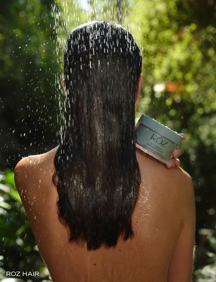 Back of model's head with wet hair holding a container of Roz scalp scrub on her shoulder.