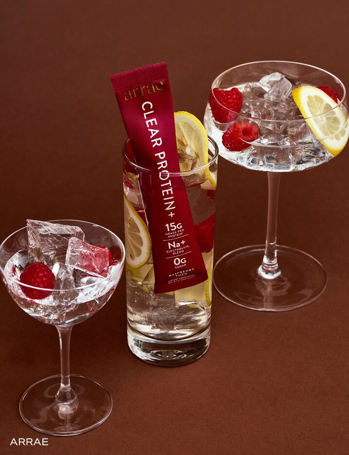 Arrae Clear Protein+ placed in a glass of iced water with sliced lemons and raspberries alongside  cocktail glasses of water.