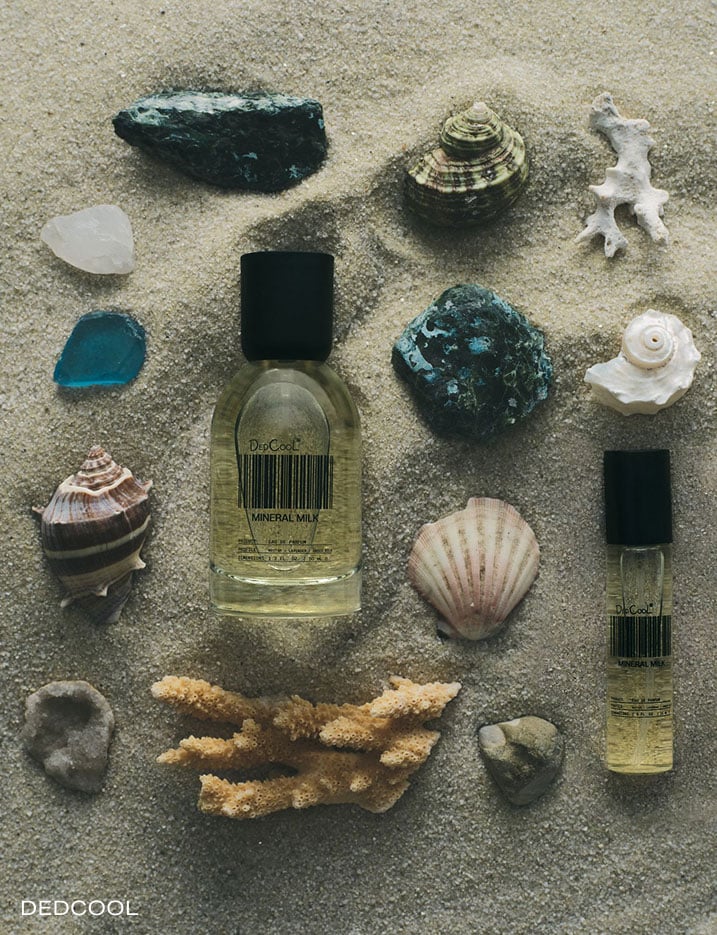 A bottle of Dedcool Mineral Milk fragrance in the sand with seashells.