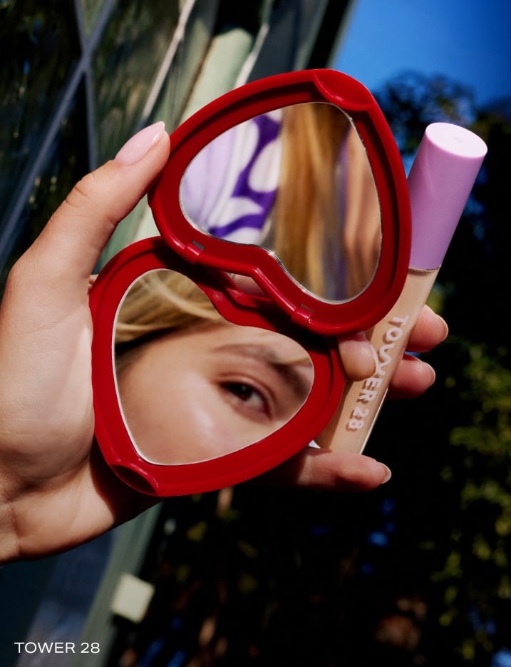 Model holding a heart shaped mirror and Tower28 Concealer reflecting her eye on the mirror.