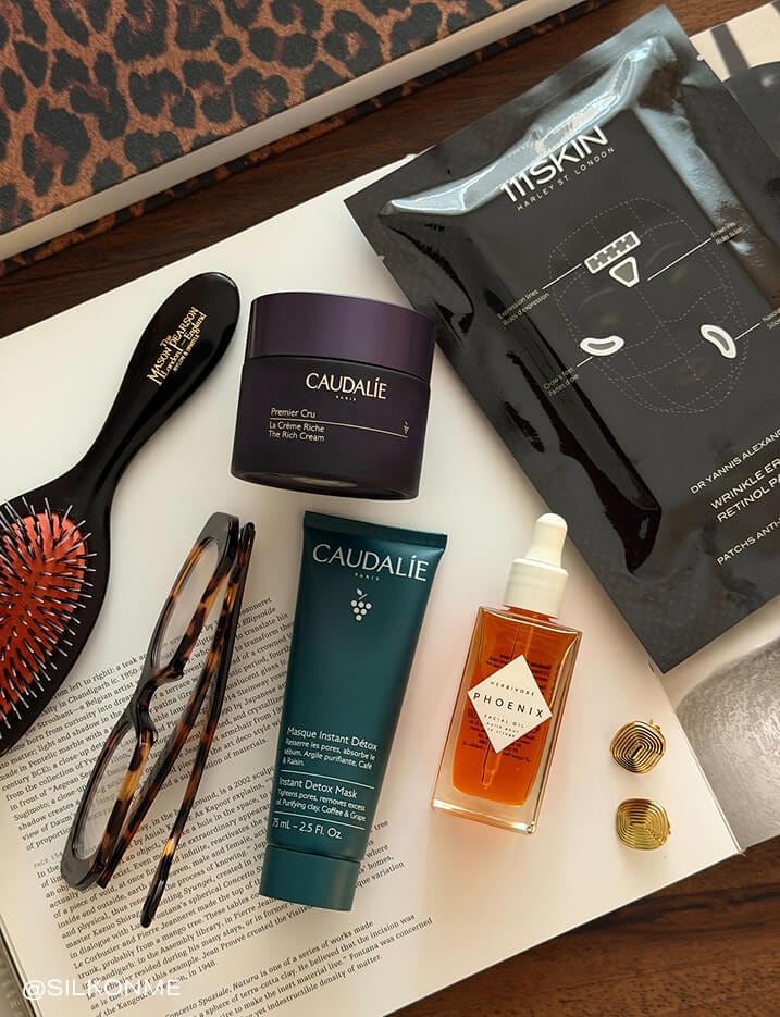 Variety of skincare alongside a brush, eyeglasses, and earrings on top of a book.