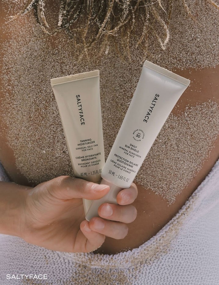Model holding SaltyFace Tanning Moisturizer and Daily Sun Shield behind her back covered in sand.